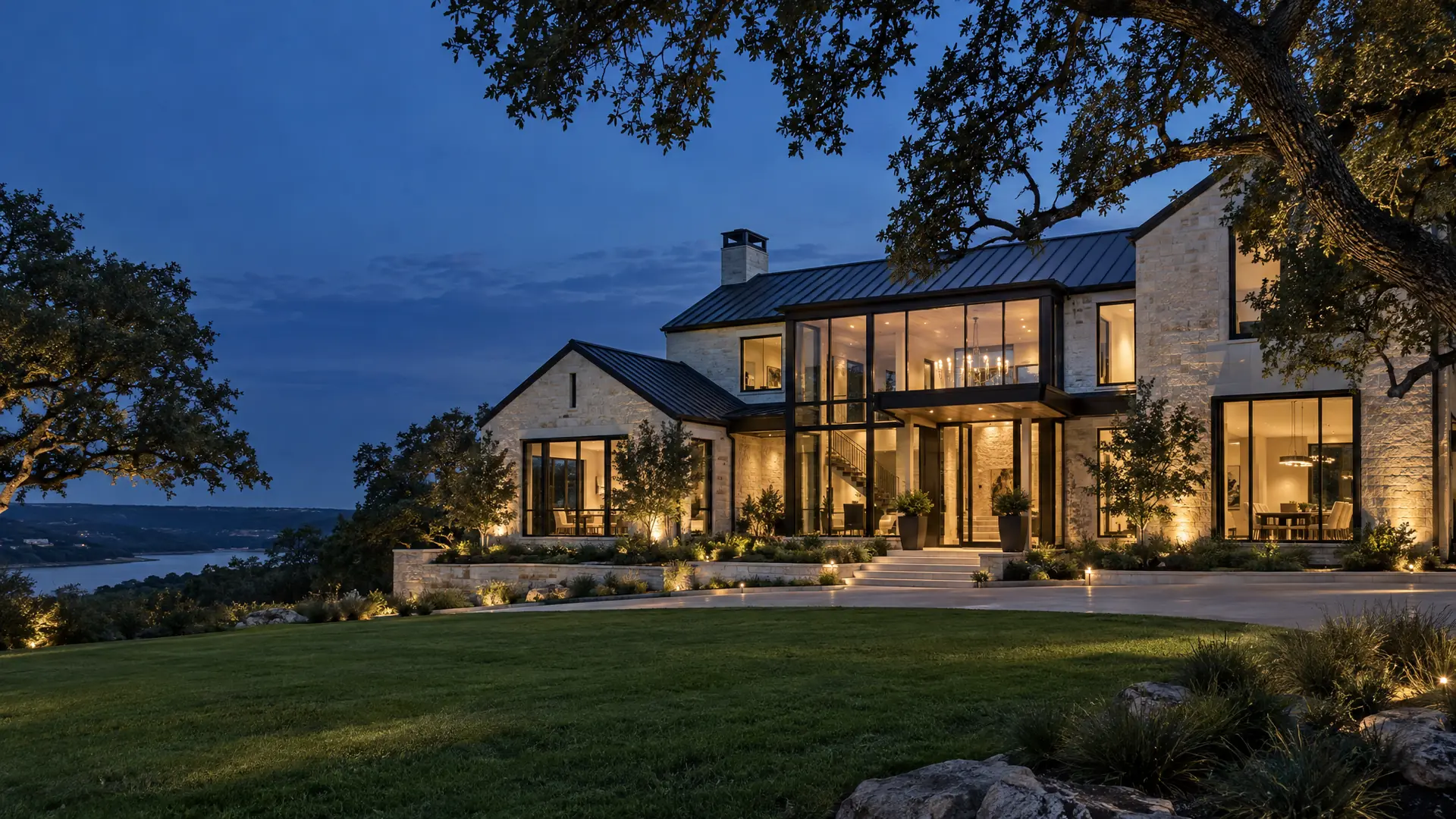 Modern luxury Texas custom home with limestone walls, metal roof and glass walls at dusk under live oaks